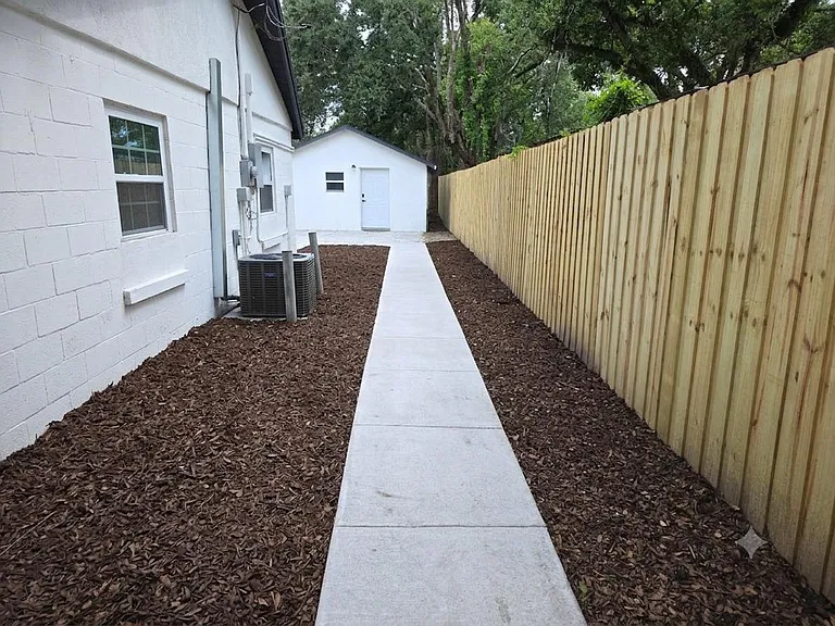 Private side yard with a concrete walkway, fresh mulch beds, and a tall privacy fence leading to a detached outbuilding.