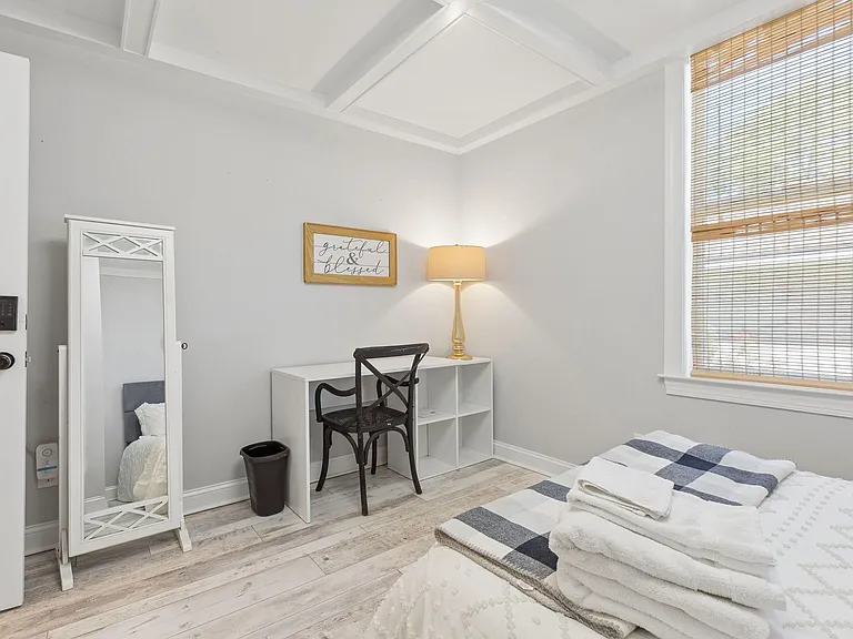 Bedroom includes a work desk, large window with blinds, and light flooring with space for a full bed and storage.