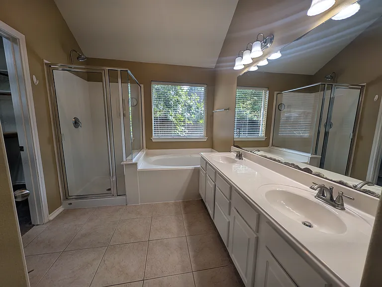 Featuring soaring vaulted ceilings, this spacious primary bath includes a long double-sink vanity, a glass-enclosed walk-in shower, and a deep soaking tub set beneath a bright window.