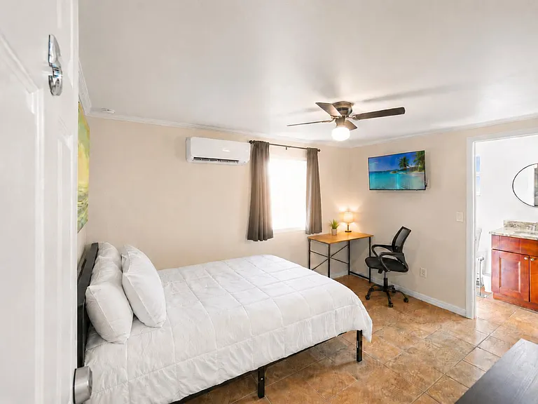 Furnished bedroom with a full bed, work desk, wall-mounted TV, ceiling fan, and window with curtains; adjacent bath access.