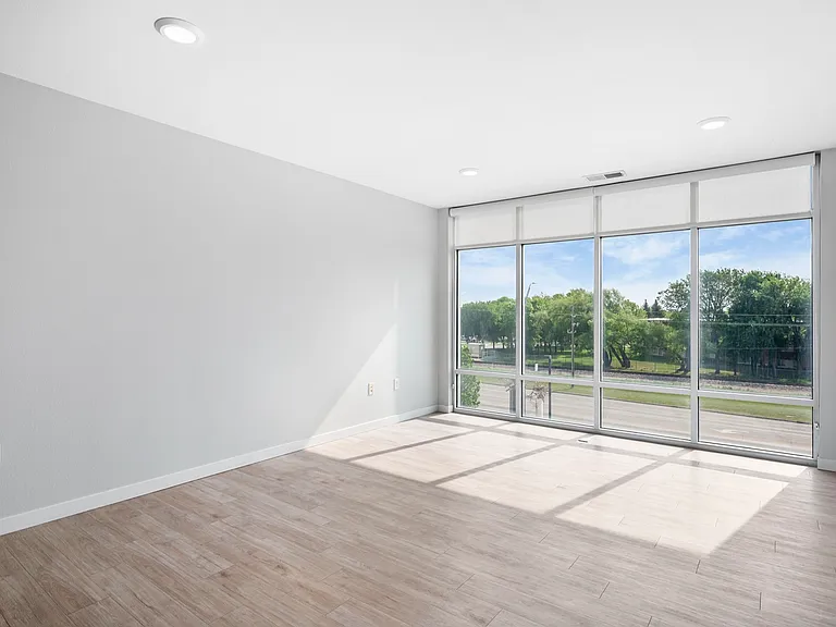 Bright and airy apartment room with large floor-to-ceiling windows offering a scenic view of greenery, complemented by sleek wood flooring and modern recessed lighting. Bright and airy apartment room with large floor-to-ceiling windows offering a scenic v