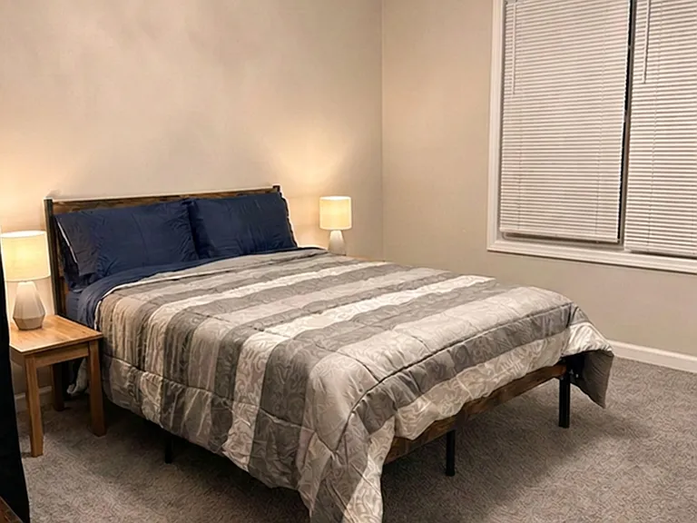 Furnished bedroom with a full bed, matching nightstands, carpet flooring, and a window with blinds for privacy.