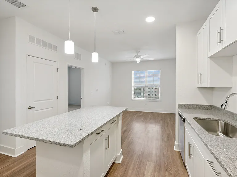 Bright, modern kitchen with sleek granite countertops and elegant white cabinetry, opening into a spacious living area with wood flooring and large windows.