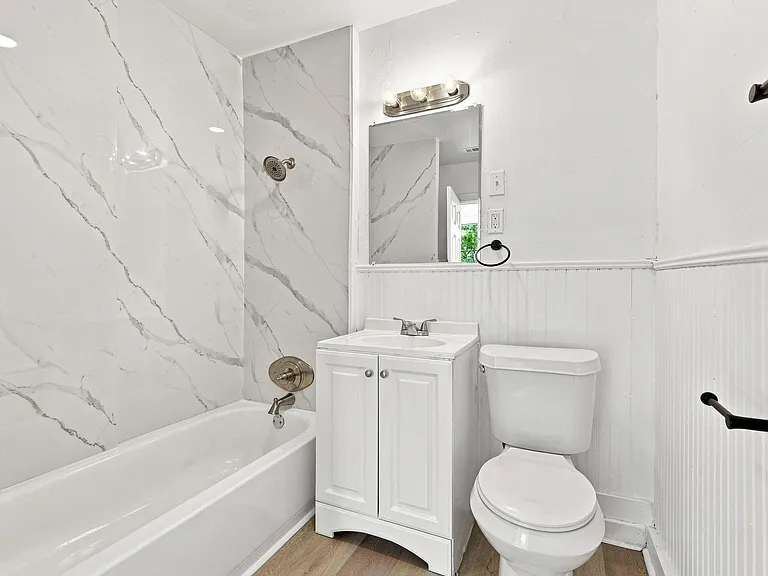 Bathroom featuring a white vanity with integrated sink, white tub with brushed nickel fixtures, marble-look shower surround, beadboard wall paneling, and wood-finish flooring