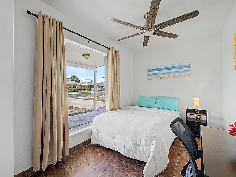 Private bedroom with large window, blackout curtains, ceiling fan, tile flooring, and space for a desk or nightstand.
