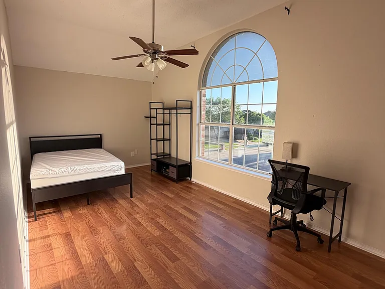 Spacious bedroom with hardwood floors, ceiling fan, large arched window, and a work desk plus open shelving/storage.