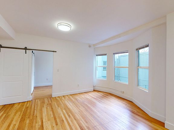 Spacious room with wooden floors and large windows letting in natural light. A sliding barn door leads to another room, creating an open feel.