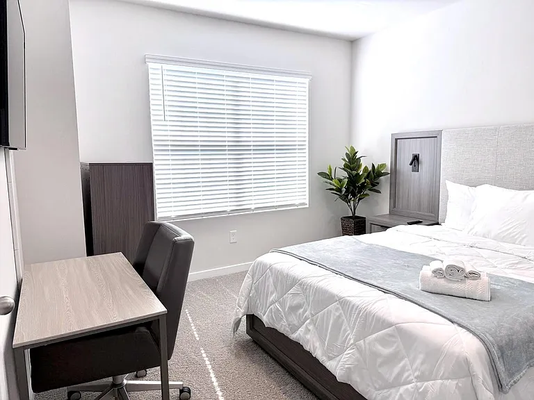 Furnished bedroom with a full bed, work desk and chair, and a wide window with blinds for natural light.