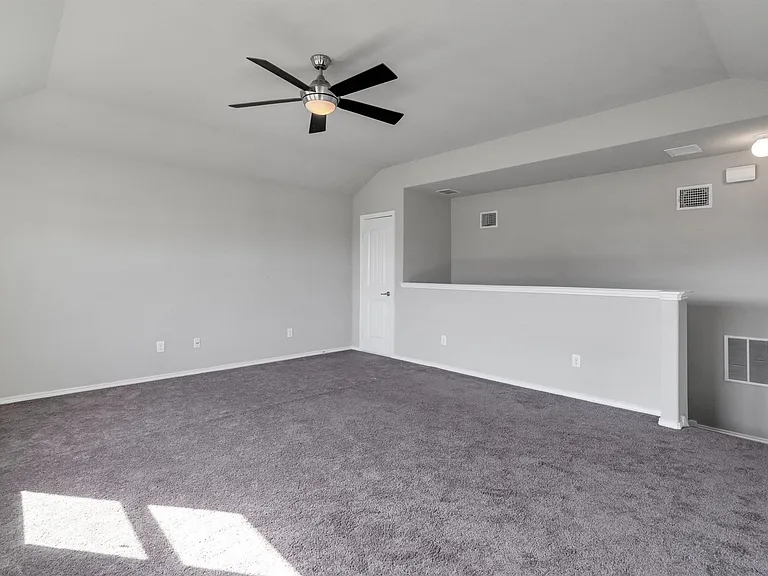Spacious room featuring vaulted ceilings, neutral gray walls, dark gray carpet flooring, a ceiling fan with integrated lighting, and a white interior door