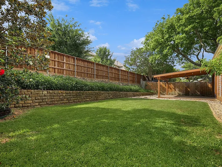 Spacious backyard featuring a manicured lawn, a retaining wall with mature landscaping, a wood privacy fence, and a covered patio area