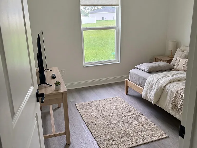 Furnished bedroom with a comfortable bed, TV setup, and a large window overlooking the yard for added natural light.