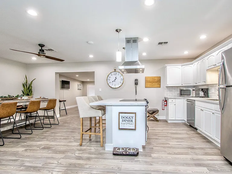 Modern kitchen at Casa Del Sol, Tempe rental home. Features bright white cabinets, stainless steel appliances, a large island with a breakfast bar, subway tile backsplash, and a pet-friendly feeding station. Ideal for a top Tempe, AZ stay.