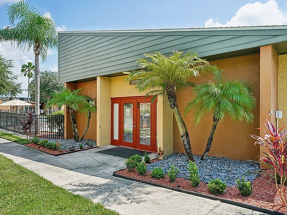 Charming and vibrant apartment entrance with tropical landscaping, featuring lush palm trees, colorful plants, and welcoming double doors.