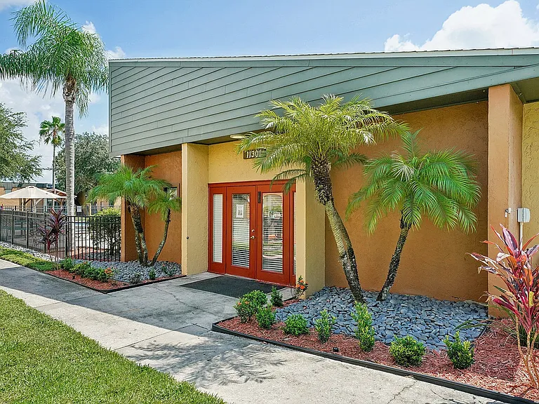 Charming and vibrant apartment entrance with tropical landscaping, featuring lush palm trees, colorful plants, and welcoming double doors.