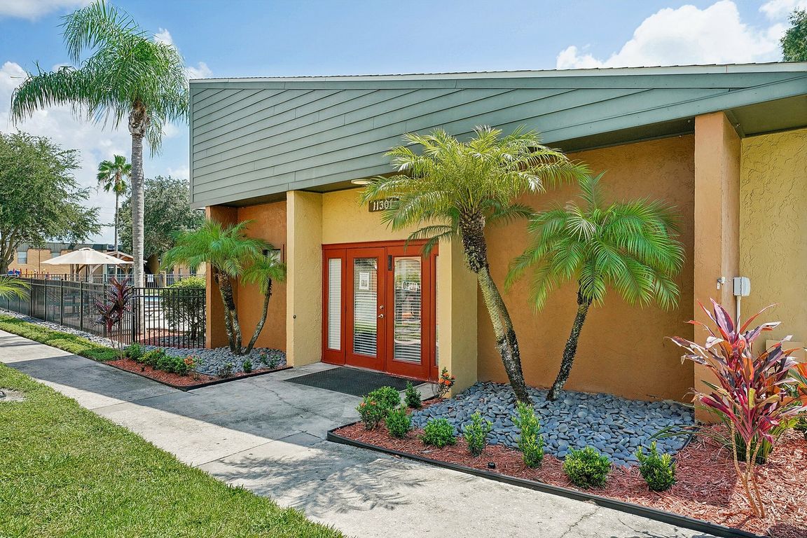 Charming and vibrant apartment entrance with tropical landscaping, featuring lush palm trees, colorful plants, and welcoming double doors.