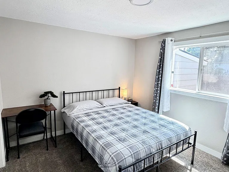 Furnished bedroom with a full bed, dedicated desk space, and a large window with curtains for natural light.