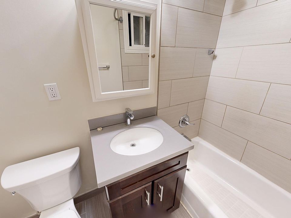 A modern bathroom with neutral tones features a toilet, a sleek vanity with a sink, a small mirror, and a bathtub with tiled walls. Bright and clean ambiance.