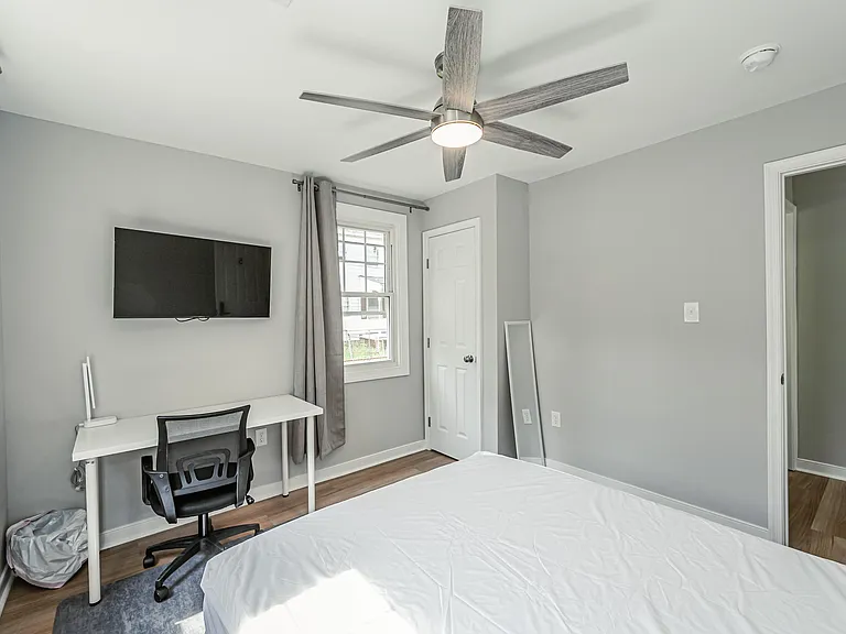 Well-appointed bedroom featuring a ceiling fan, wall-mounted TV, dedicated desk space, and a window with curtains.