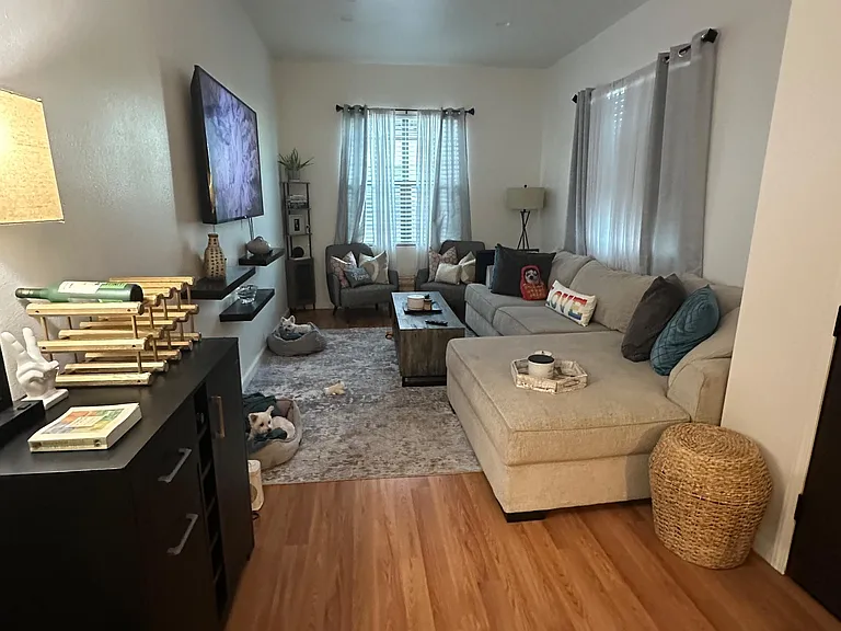 Spacious living room with sectional seating, wall-mounted TV, and large windows with curtains over hardwood floors.