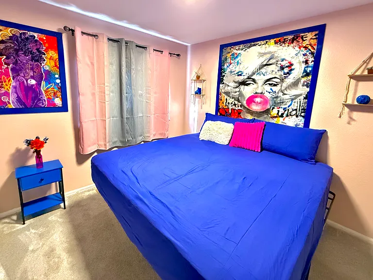 The bedroom exudes vibrant & eclectic energy with its bold, colorful artwork & furnishings. The large bed, adorned with deep blue linens, invites relaxation, while the bright artwork adds a playful touch. Natural light streams through the window.