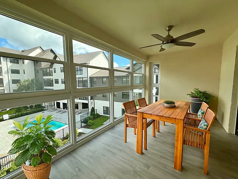 Bright and airy apartment balcony with elegant wooden dining set, large windows, and a scenic view of the courtyard pool and modern apartments.