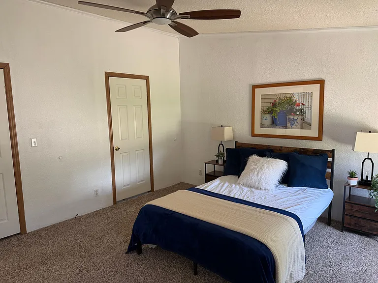 Furnished bedroom with a ceiling fan, carpet flooring, and a comfortable bed flanked by matching nightstands and lamps.