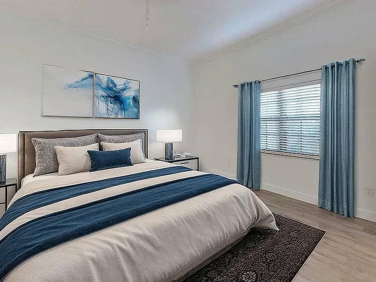 Inviting bedroom with elegant decor, a plush bed adorned with blue accents, and ample natural light from a large window with stylish curtains.
