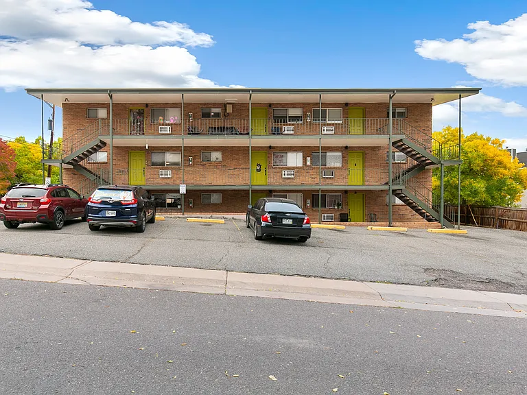 Charming two-story apartment building with vibrant green doors, ample parking, and a picturesque backdrop of autumn foliage.