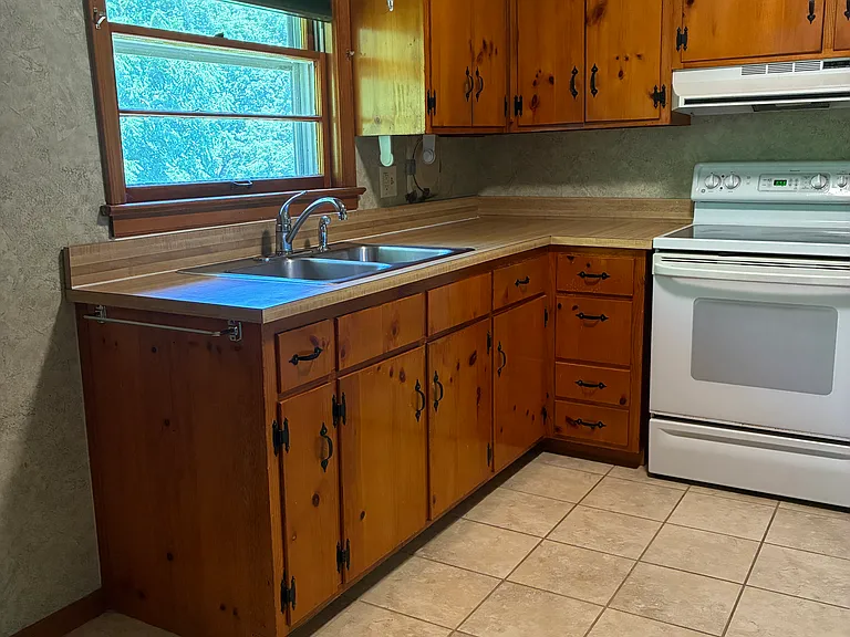 Bright, classic kitchen with warm wood cabinets, full-size range, refrigerator/freezer, beautiful tile flooring, and natural light from the window over the sink.