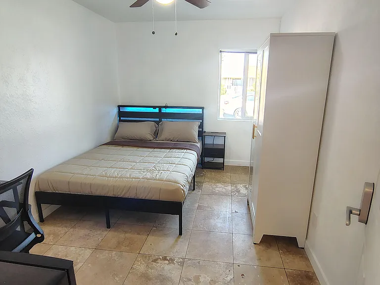 Furnished bedroom with a ceiling fan, tiled floors, a large wardrobe, and a window for natural light.
