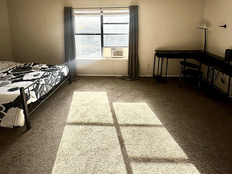Carpeted bedroom with a large window, curtain panels, in-room A/C, and space for a bed plus a work desk.