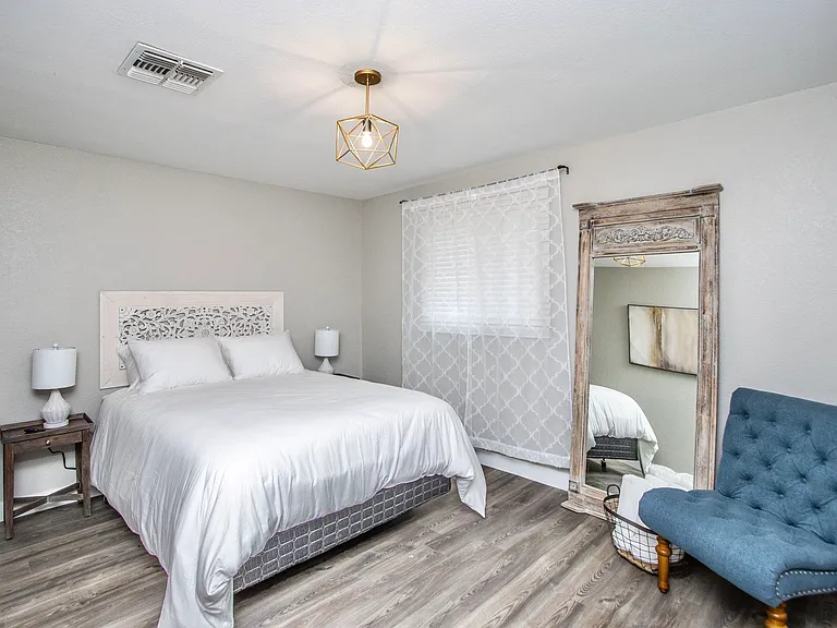 Serene primary bedroom at Casa Del Sol, Tempe rental home. Features a comfortable bed with a carved wood headboard, a large rustic full-length mirror, a blue accent chair, and modern geometric lighting. A peaceful Tempe, AZ vacation retreat.