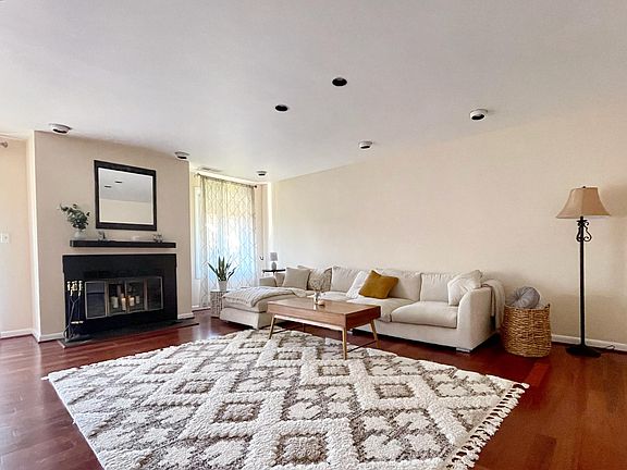 Spacious well-lit living room with white couch, beautiful rug, coffee table, fire place / mantel, and large windows overlooking the neighborhood gardens.