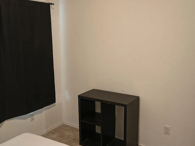 Private bedroom with carpeted flooring, blackout window covering, and a compact shelving unit for convenient storage.