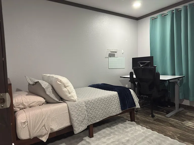 Furnished bedroom with a full bed, spacious work desk, and window with blackout-style curtains for privacy and comfort.