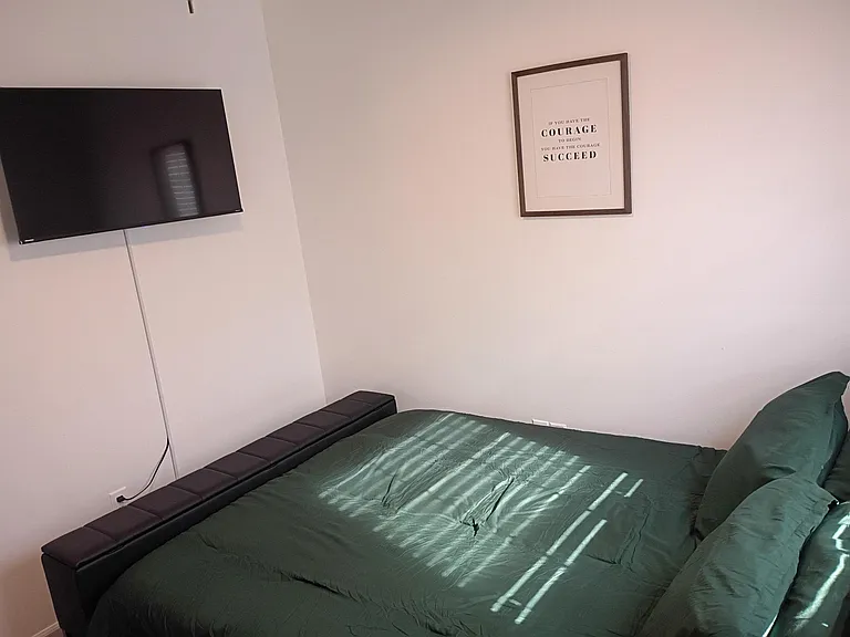 Furnished bedroom with a queen bed, ceiling fan, wall-mounted TV, and window blinds for light and privacy control.