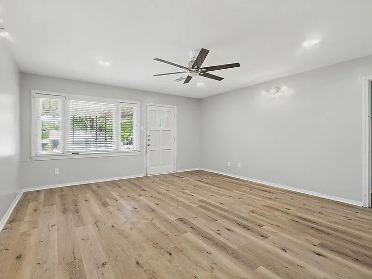 Spacious living room with neutral soft grey paint, LVP flooring, recessing lighting, a ceiling fan and ample amount of light from the large window.