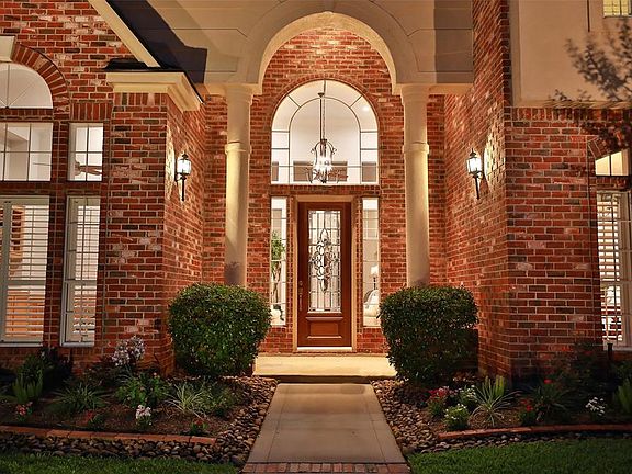 AN AMAZING VIEW OF THE ENTRY AT DUSK WITH AN 8 FOOT DOOR AND IONIC COLUMNS FLANKING THE ARCHED FRONT PORCH - Enter this remarkable home into a two-story grand entry revealing hardwood floors and a drop down iron chandelier with recessed lighting - vi
