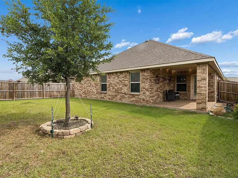 Expansive backyard featuring a covered patio, full brick exterior, privacy fencing, and a mature tree with a stone-edged planting bed