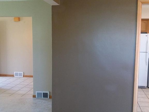 Open floor plan view from dining room. In photo from left to right is carpeted living room, tile entry, kitchen. All paint is a shade of beige/brown.