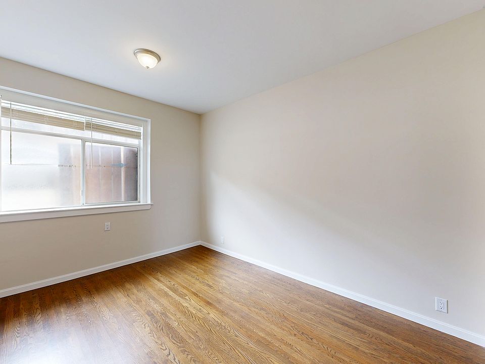 Bright, empty room with wood flooring, lit by a window on the left. A ceiling light fixture adds warmth, enhancing a cozy ambiance.