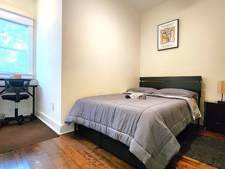 Furnished bedroom with full-size bed, nightstand, and a dedicated desk area near the window for work or study.