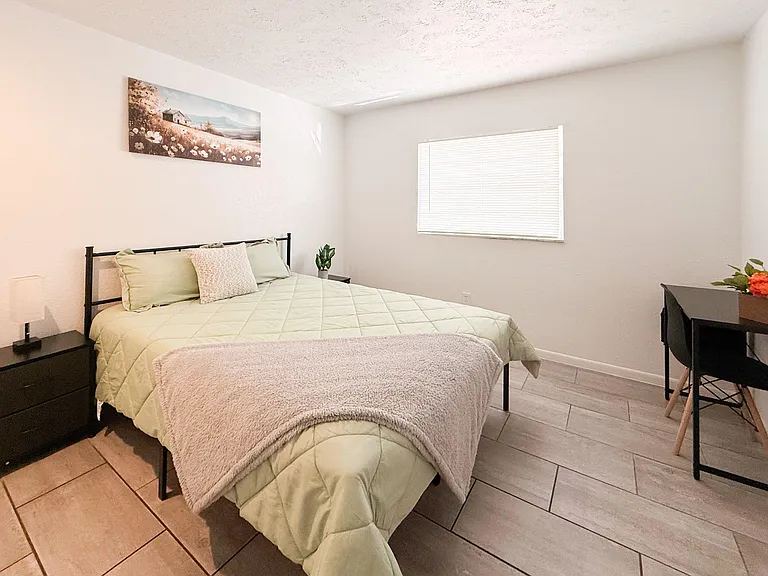 Furnished bedroom with a full bed, nightstand, and desk area, plus a window with blinds and tile flooring.