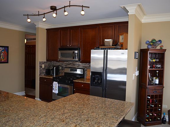 Kitchen with stainless steel appliances and modern lighting. Large counter can accommodate 4 counter stools. Rich tile back splash above new granite counter tops. Kitchen has a dishwasher + disposal. Kitchen floor is tiled; entry hall in background h