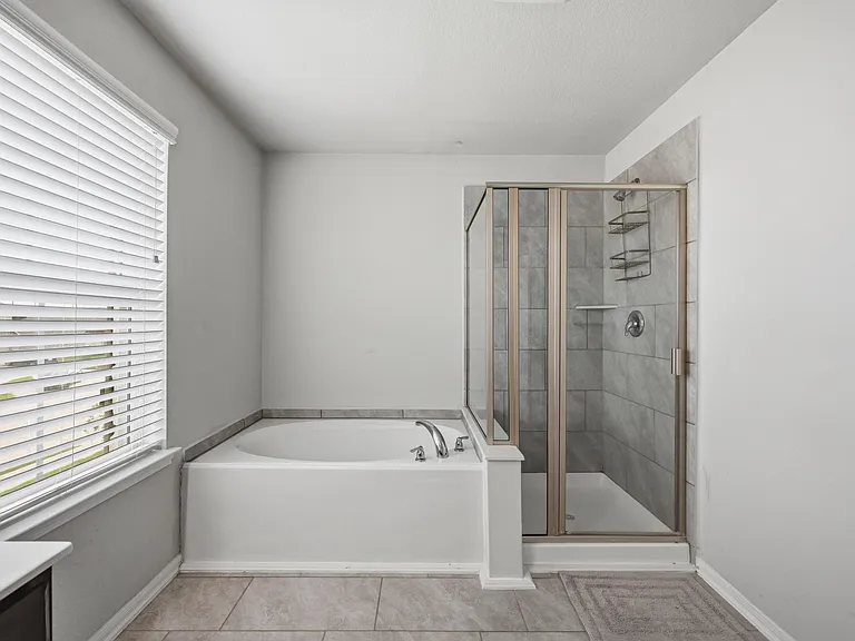Spacious bathroom featuring a soaking tub, separate glass-enclosed tile shower, and a large window with blinds.
