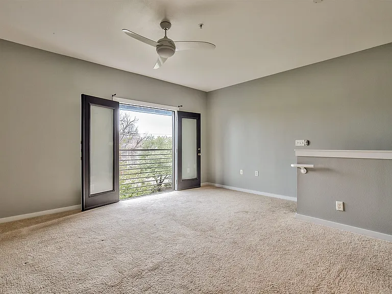 Room with light gray walls and beige carpeting, featuring double doors opening to a balcony with a metal railing