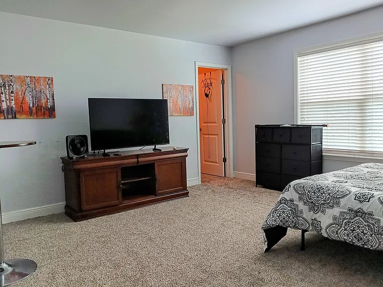 Spacious carpeted bedroom with a large window and blinds, plus a dresser and TV stand for convenient storage and setup.