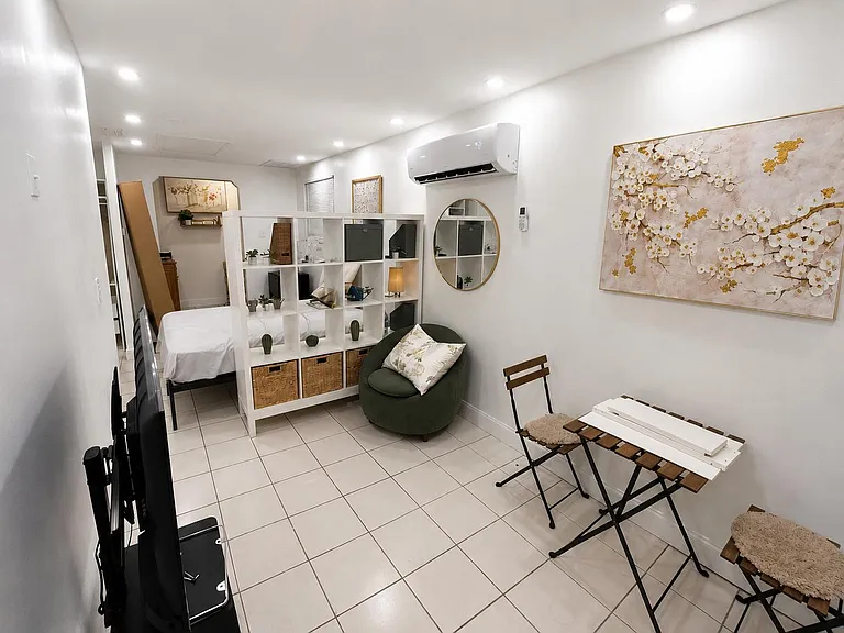 Flexible studio-style sleeping area with tile floors, room divider shelving, and a dedicated sitting nook under wall AC.