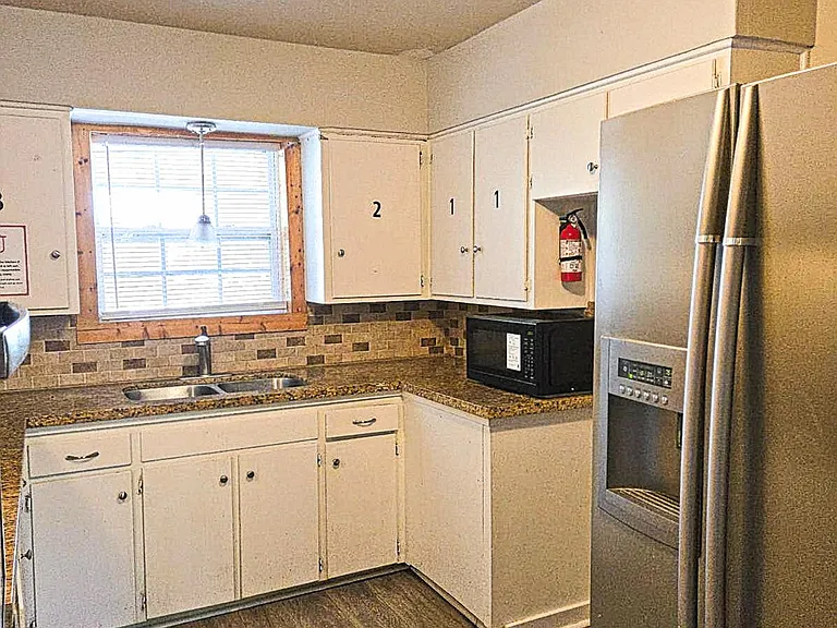 Compact L-shaped kitchen with ample cabinetry, double-basin sink, tile backsplash, and stainless side-by-side fridge.