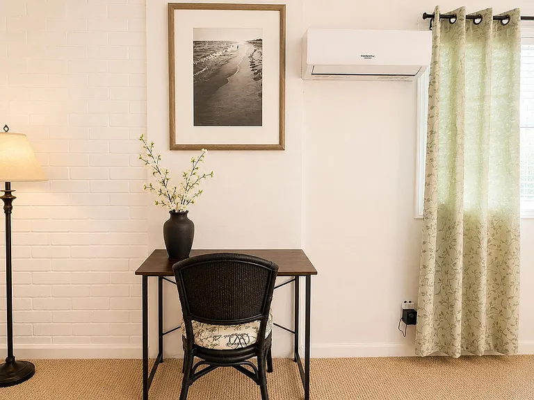 Quiet work nook with a writing desk, chair, wall art, and a nearby window with curtains plus mini-split A/C.